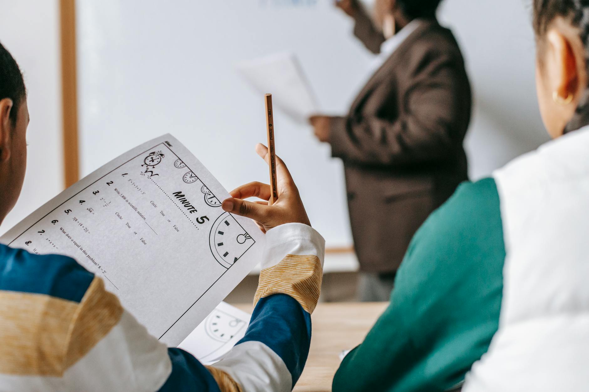 Students in a classroom focus on worksheets while the teacher conducts a lesson.