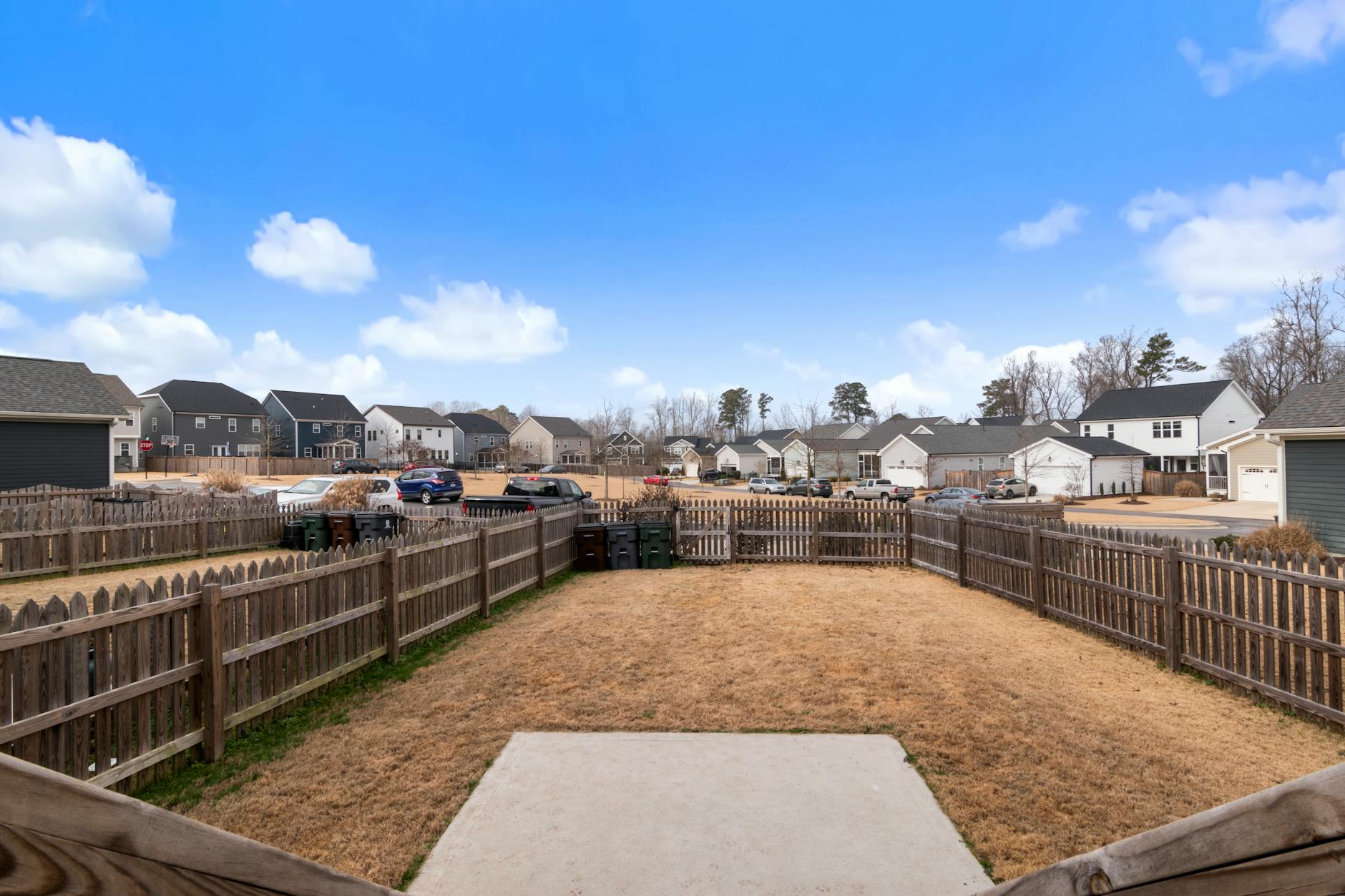 A suburban backyard with wooden fencing and modern houses under a blue sky.